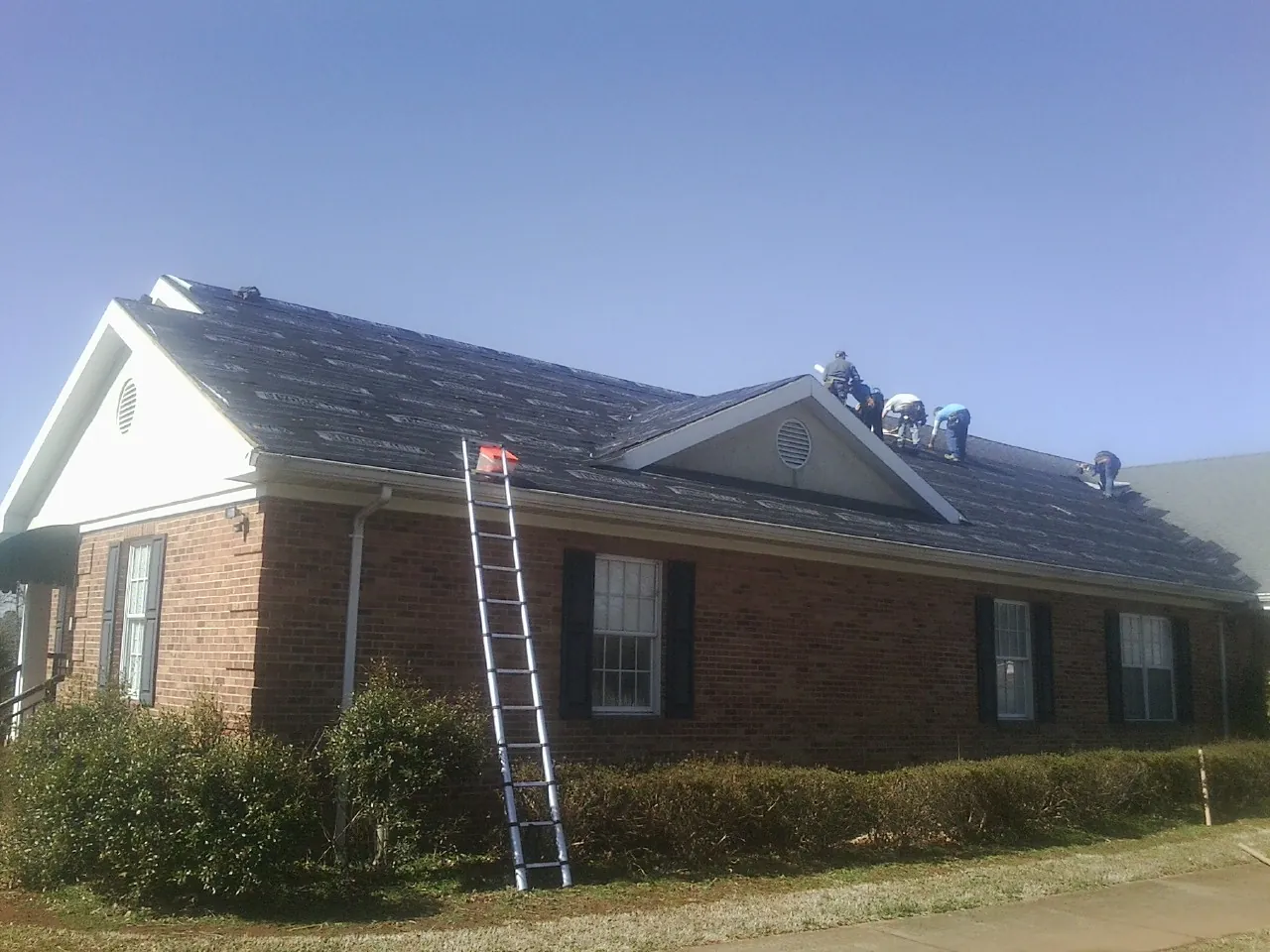 Crew tearing shingles off a brick ranch roof with a ladder set — Emergency Roof Repair work by our crew in Cranford