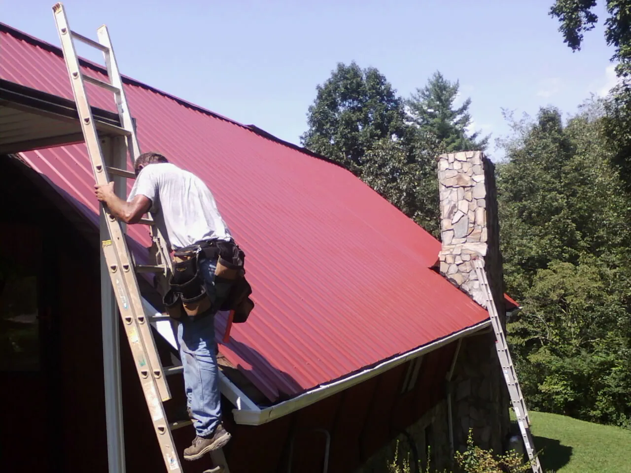 Roofer on a ladder beside a red metal roof and stone chimney — Soffit & Fascia Repair by Roofing Brothers in Cranford